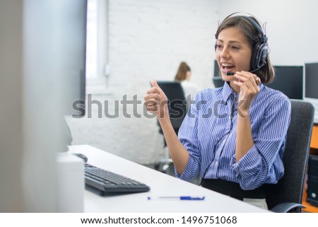 Beautiful friendly female costumer service operator showing thumb up to computer screen during video call in call centre