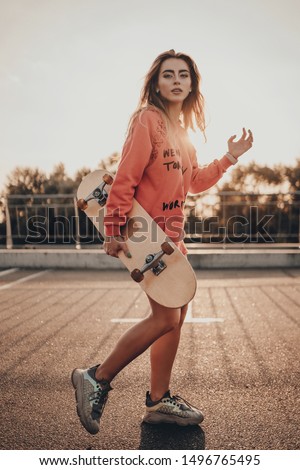 Urban stylish young girl walking with skateboard in red hoodie and sneakers on summer sunset on parking near road. Outdoors portrait of active sport woman model Royalty-Free Stock Photo #1496765495