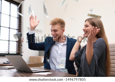 Happy young people playing online lottery using laptop under money rain in office