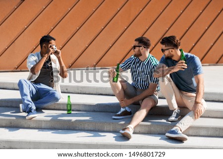 leisure, male friendship and people concept - man with camera photographing his friends drinking beer on street in summer