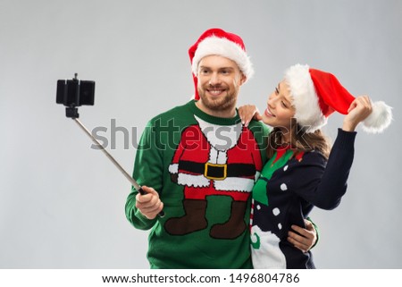 christmas, technology and holidays concept - happy couple in santa hats taking picture by smartphone on selfie stick at ugly sweater party