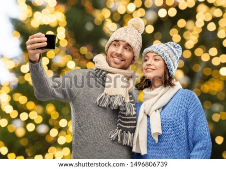 people, christmas and winter clothes concept - happy couple in knitted hats and scarves taking selfie by smartphone over festive lights background