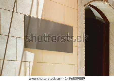 Perspective view of a modern contemporary big metallic name plate with copy space on a nice white historical stone wall with areas of shade and rays of natural sunlight and next to a wooden door.