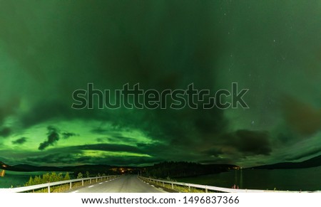 Northern Lights panoramic photo of strong bright Aurora Borealis behind heavy clouds over road, Joesjo Lake and Scandinavian mountains, Swedish Lapland look very dramatic, Northern Sweden. Late summer