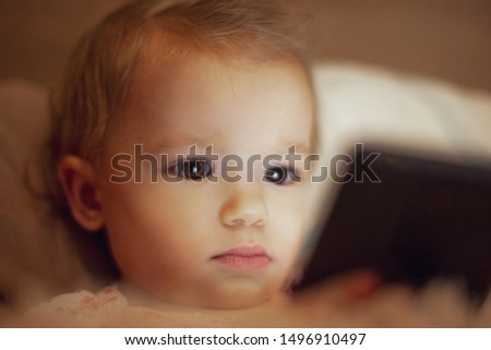 a little girl lying in bed in a room with the light off and holding a smartphone, playing games and watching cartoons. Baby addiction to gadgets