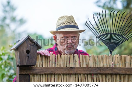 An elderly man with hat looks angry and watching over a garden fence. Concept problems with the neighborhood. Royalty-Free Stock Photo #1497083774