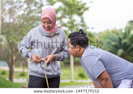Two people, muslim woman and fat man, proportional and over weight, overweight asian male look envy at his female muslim woman slim body at outdoor city park