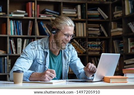 Smiling happy young man wear wireless headset look at laptop screen make notes study e learning in library watch webinar training online course video call, distance education, skype teaching concept Royalty-Free Stock Photo #1497205910