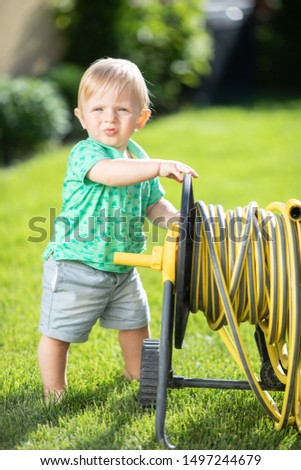 one year child outdoor with green grass background. little blond baby boy standing on the backyard with hose for watering the lawn. baby boy on the grass. child on summer time. little helper watering 