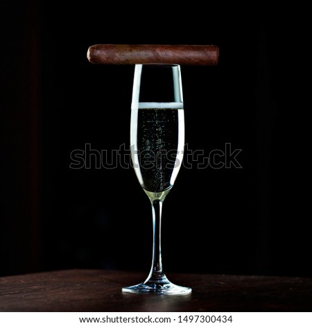 Picture of tall glass of champagne and Cuban cigar on top of it on wooden   table with black background