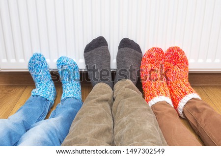 Mother, father and son wearing colorful of pair woolly socks warming cold feet in front of heating radiator at home. The symbolic image of the heating season at home. Part of body, selective focus. Royalty-Free Stock Photo #1497302549