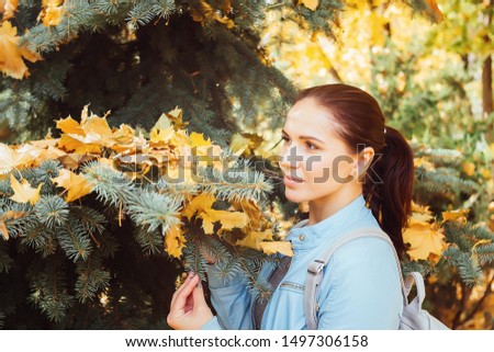 Blindfolded brunette girl in a blue leather jacket looks at the yellow fallen maple leaves on the branches of spruce. She exposes her face to the sun. Soft picture.