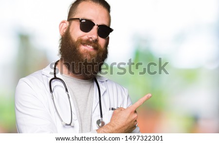 Young blond doctor man with beard wearing sunglasses with a big smile on face, pointing with hand and finger to the side looking at the camera.