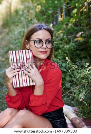 
Student, sitting on the blanket in park, holding striped notebook, thinking. Close-up picture of young blond woman, wearing eyeglasses, red blouse, studying at nature, College campus in summer.
