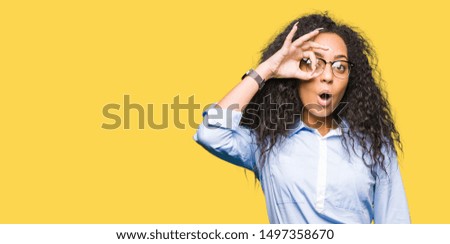 Young beautiful business girl with curly hair wearing glasses doing ok gesture shocked with surprised face, eye looking through fingers. Unbelieving expression.