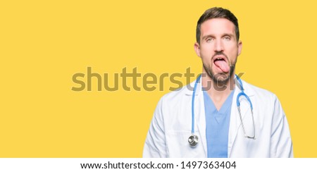 Handsome doctor man wearing medical uniform over isolated background sticking tongue out happy with funny expression. Emotion concept.