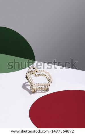 Detail close-up shot of a heart-shaped hair claw decorated with a quantity of shining pearls. The golden hair clip is isolated in the light interior with red and green geometric details.