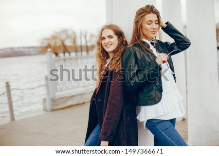 a beautiful stylish young girl with long curly hair and a long coat standing in a autumn park with her girlfriend in a black leather jacket and glasses