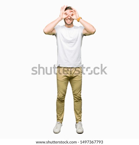Handsome man wearing casual white t-shirt doing ok gesture like binoculars sticking tongue out, eyes looking through fingers. Crazy expression.