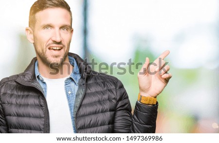Handsome man wearing winter coat with a big smile on face, pointing with hand and finger to the side looking at the camera.