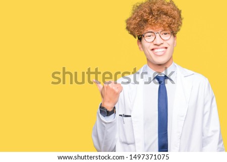 Young handsome scientist man wearing glasses smiling with happy face looking and pointing to the side with thumb up.