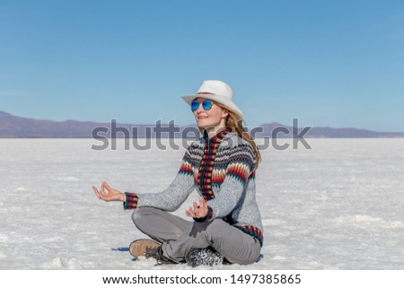 Cheerful positive blond girl clothing casual sitting at the salt flat desert Uyuni and meditating in yoga pose. Caucasian casual model wearing hat and sunglasses. Landscape background with copy space