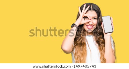 Young adult woman showing smartphone screen with happy face smiling doing ok sign with hand on eye looking through fingers