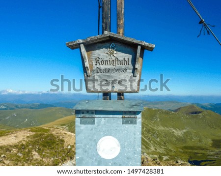 A close-up on the middle part of a cross, on which there is a sigh saying:Königstuhl, Dreiländereck 2330m, and a metal box. In the back there are endless chains of mountains. Clear and sunny day.