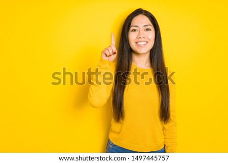 Beautiful brunette woman over yellow isolated background showing and pointing up with finger number one while smiling confident and happy.