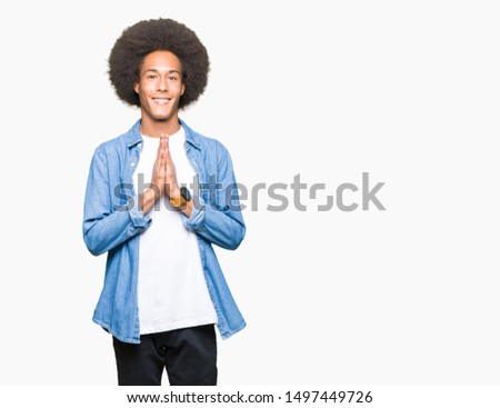 Young african american man with afro hair praying with hands together asking for forgiveness smiling confident.