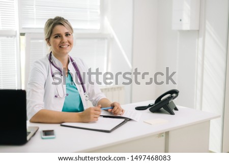 Portrait of young smiling woman doctor in hospital office with laptop taking patient. Female therapist sits at the table in front of the camera and looking at camera