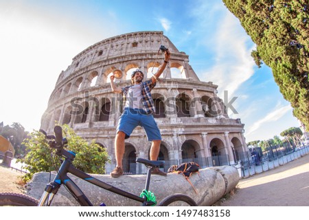 Happy young man tourist with bike wearing shirt and waving hat standing on a column taking selfies at colosseum in Rome, Italy at sunrise.