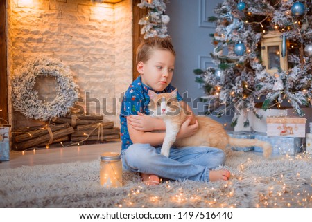 The boy sits on the floor near the fireplace and holds a red cat. Christmas concept, toy tree in the background