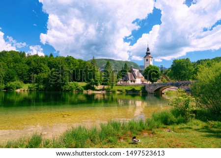 Scenery of Church of Saint John Baptist on Bohinj Lake in Slovenia. Nature in Slovenija. View of blue sky with clouds. Beautiful landscape in summer. Alpine Travel destination. Julian Alps mountains