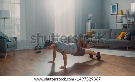 Muscular Athletic Fit Man in T-shirt and Shorts is Doing Push Up Exercises at Home in His Spacious and Sunny Living Room with Modern Interior. Royalty-Free Stock Photo #1497529367