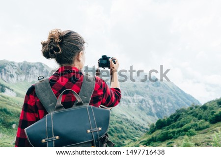 A view from the back of a female photographer, a blogger taking a photo of an epic mountain landscape. Adventure travel with backpack and camera.