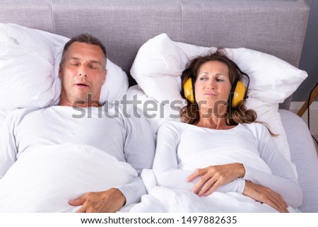 Mature Woman Covering Her Ears With Headphone While Man Snoring In Bed Royalty-Free Stock Photo #1497882635