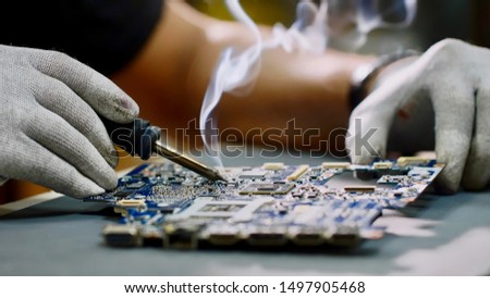 Technician engineer in workshop. Repairman in gloves is soldering circuit board of electronic device on the table, hands close up. He takes tin with a soldering iron and puts it on microcircuit. Royalty-Free Stock Photo #1497905468