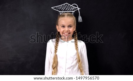 Smiling girl standing with academic cap painted on blackboard, school education