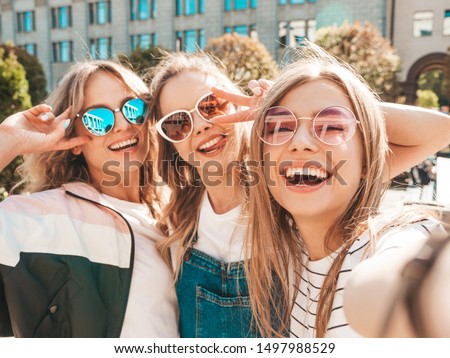 Three young smiling hipster women in summer clothes.Girls taking selfie self portrait photos on smartphone.Models posing in the street.Female showing positive face emotions in sunglasses
