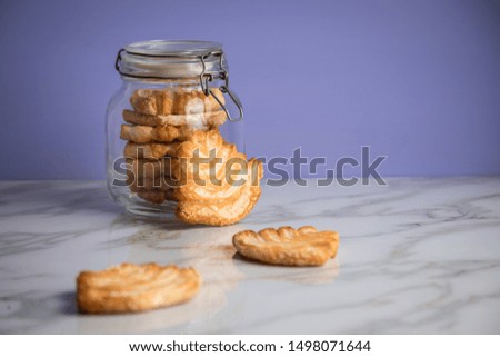 Italian puff pastry fan wavers cookies biscuits with preserving glass jar on marble table and lilac background