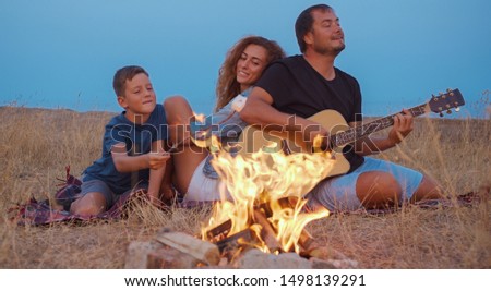 Daddy plays guitar , mom with son enjoy while relaxing on family picnic near bonfire.