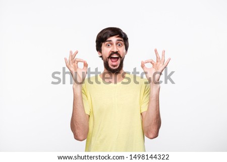 Handsome brunet bearded man with mustaches in a yellow shirt showing gesture of OK sign with two hands standing isolated over white background. OK gesture.