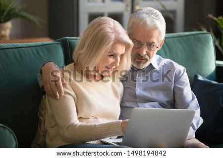 Smiling elderly grey-haired couple siting on sofa at home, looking in computer, watching family photos, resting, spending free time in living room. Attractive aged woman helping husband with new app