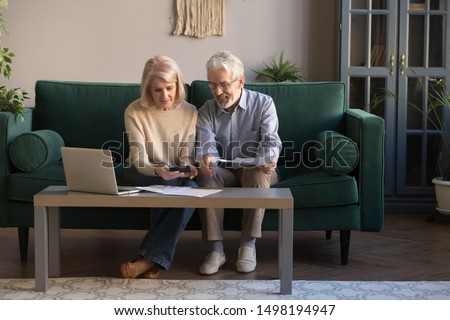 Smiling contemporary senior couple spouses use laptop new technologies paying banking bills online, happy modern mature husband and wife calculate finances, manage domestic expenditures at home Royalty-Free Stock Photo #1498194947