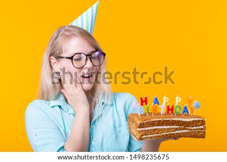 Funny positive young woman holds in her hands a homemade cake with the inscription happy birthday posing on a yellow background. Concept of holidays and anniversaries.