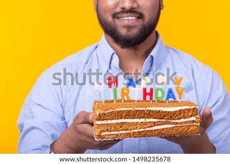 Close-up of funny young indian guy with a cap and a homemade cake in his hands posing on a yellow background. Anniversary and birthday concept.