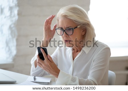 Confused senior businesswoman sit at office desk hold cellphone experience internet connection problem, frustrated aged woman worker feel disappointed having smartphone breakdown or virus attack Royalty-Free Stock Photo #1498236200