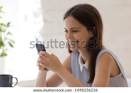 Smiling girl sit at desk feel happy texting messaging on smartphone with friends, excited young woman hold cellphone chatting or browsing internet, overjoyed female use new technology device