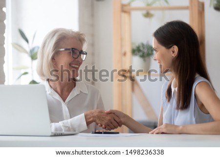 Happy female business partners get acquainted handshake near laptop at office meeting, smiling senior businesswoman shake hand greeting with millennial woman client or job applicant